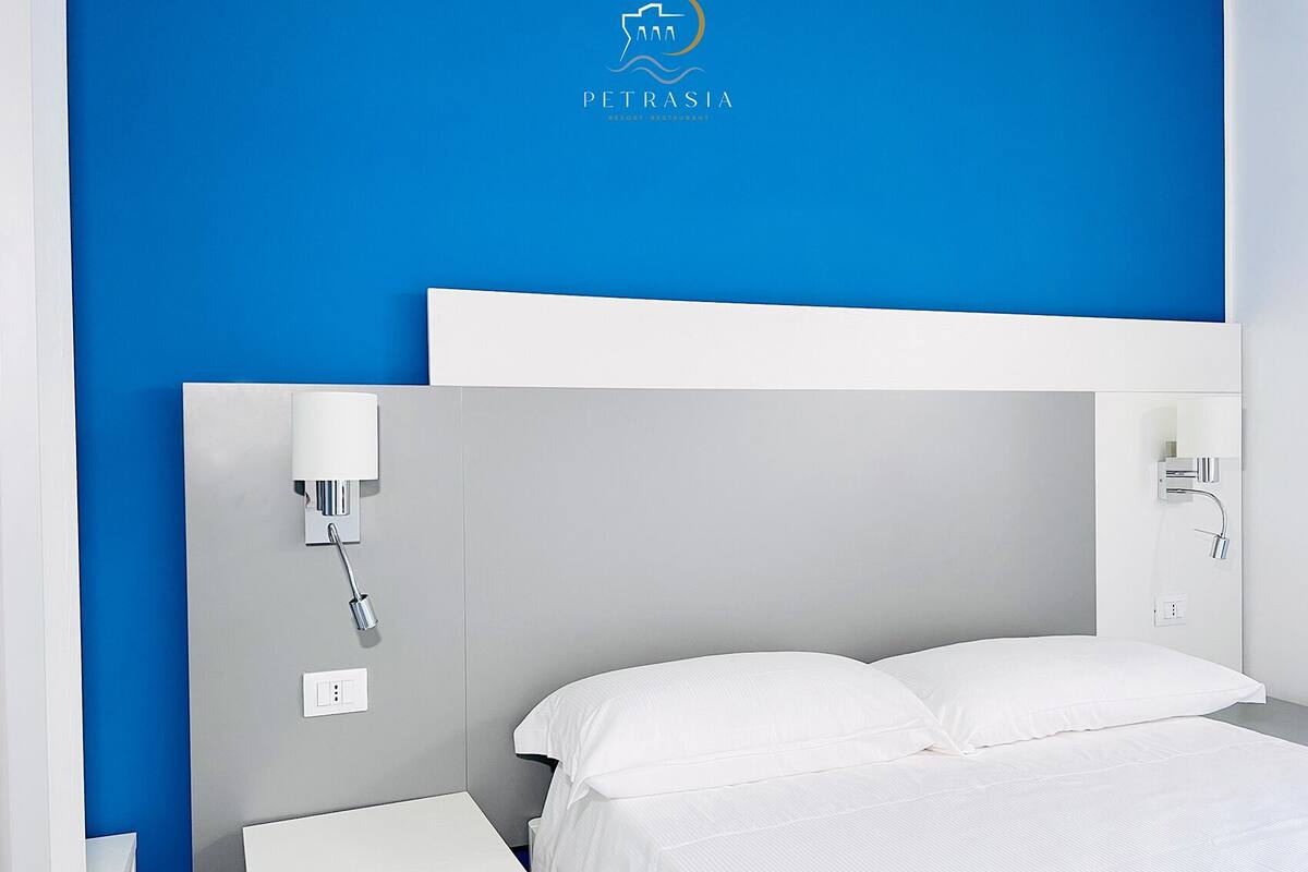 A modern hotel room with a blue accent wall, a white bed, and a grey headboard.