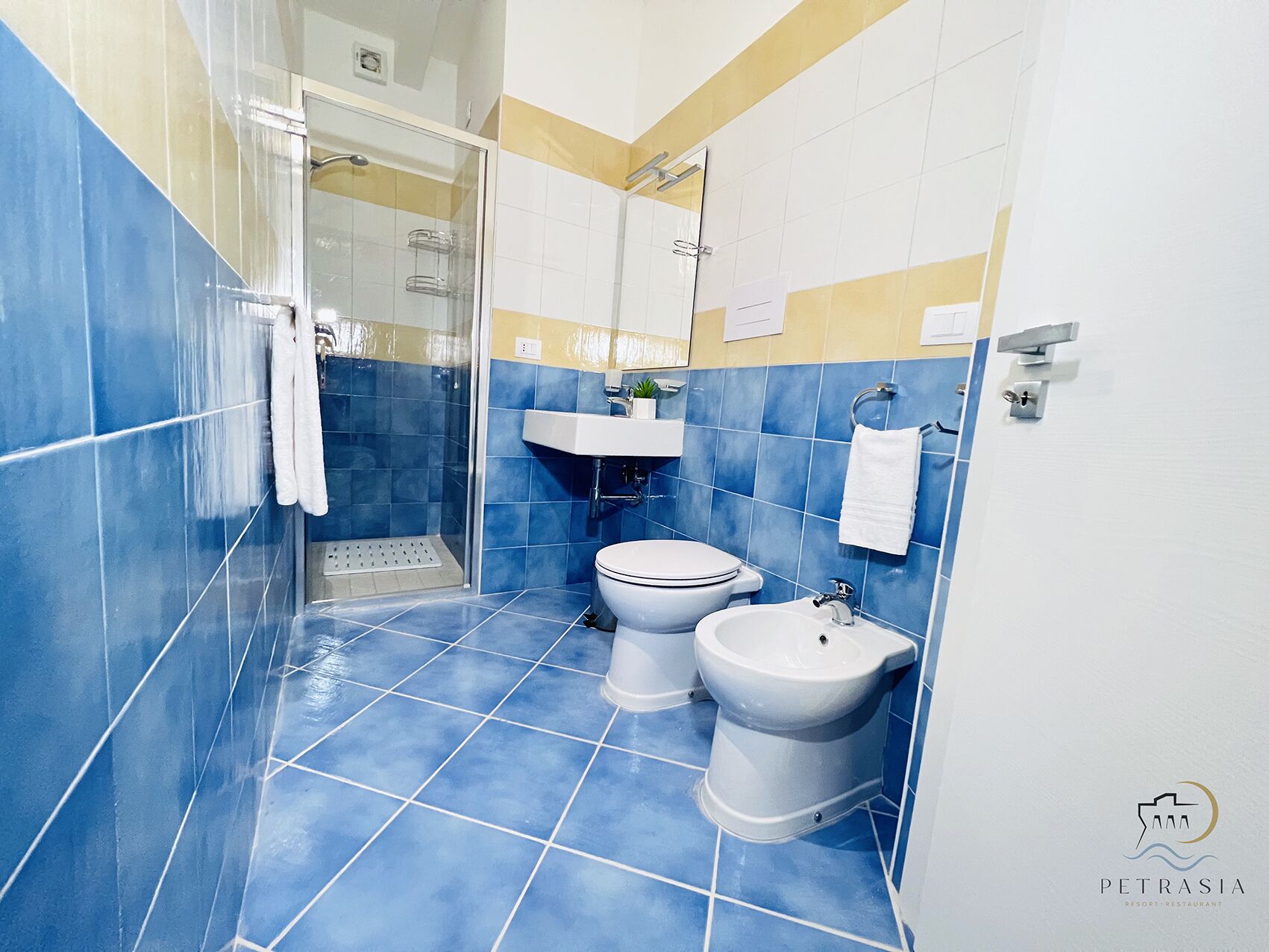 Ein Badezimmer mit blauen Fliesen, einer Duschkabine aus Glas, einer Toilette, einem Bidet und einem Waschbecken.