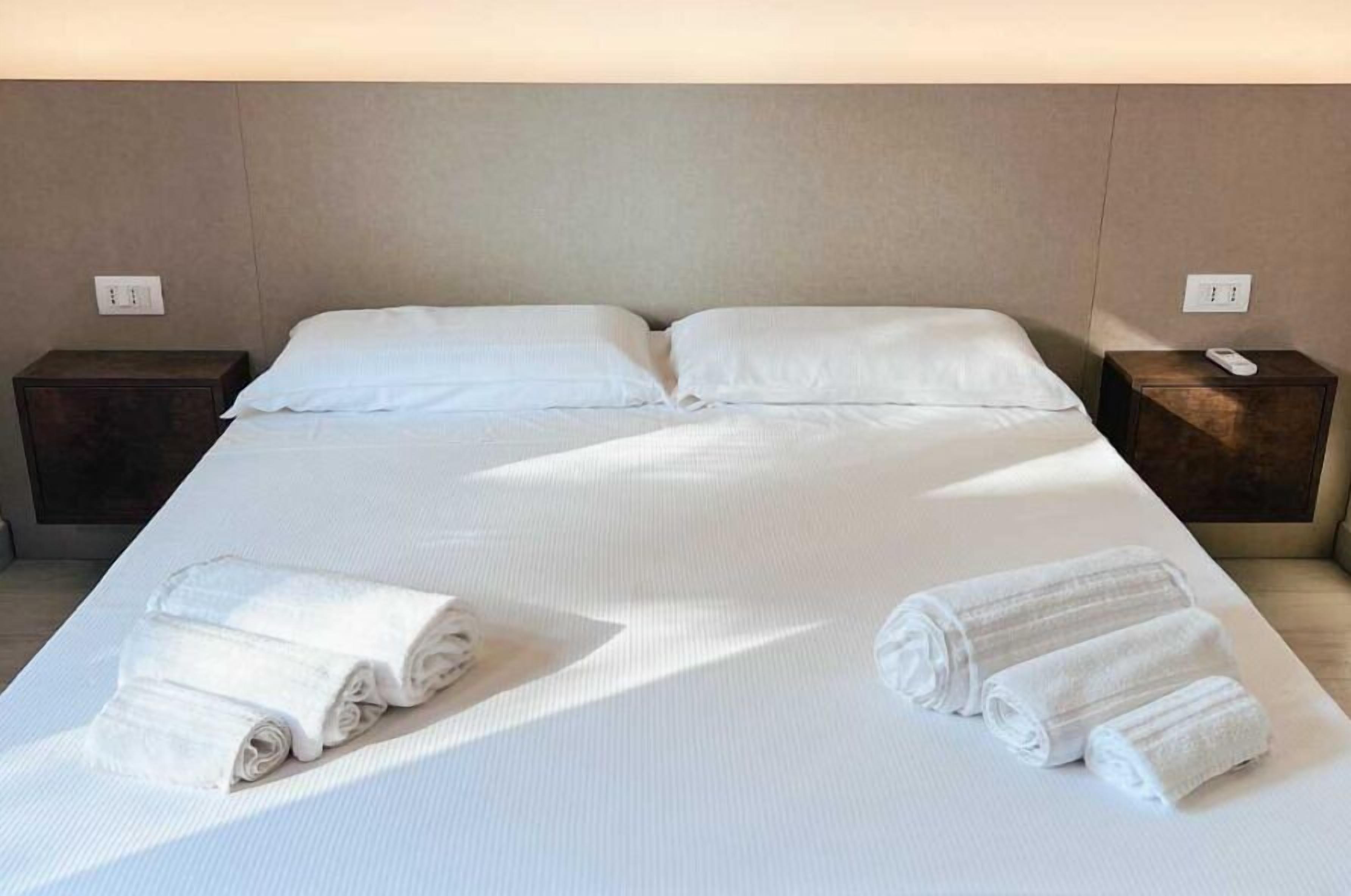 A neatly made bed with white linens, two bedside tables, and rolled towels on the bed.