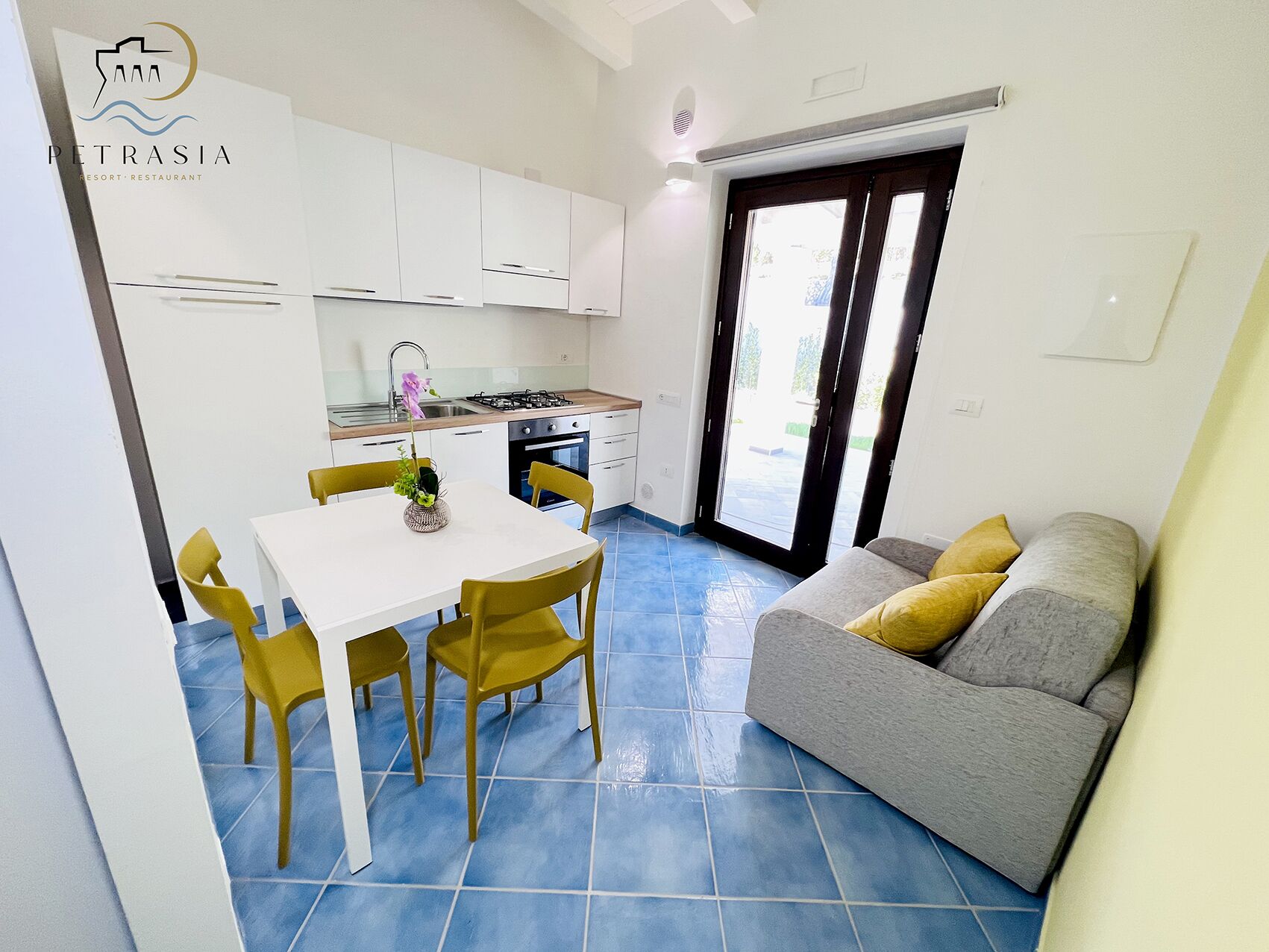 Una cucina compatta con mobili bianchi, un tavolo da pranzo con sedie gialle e un divano grigio con cuscini gialli.