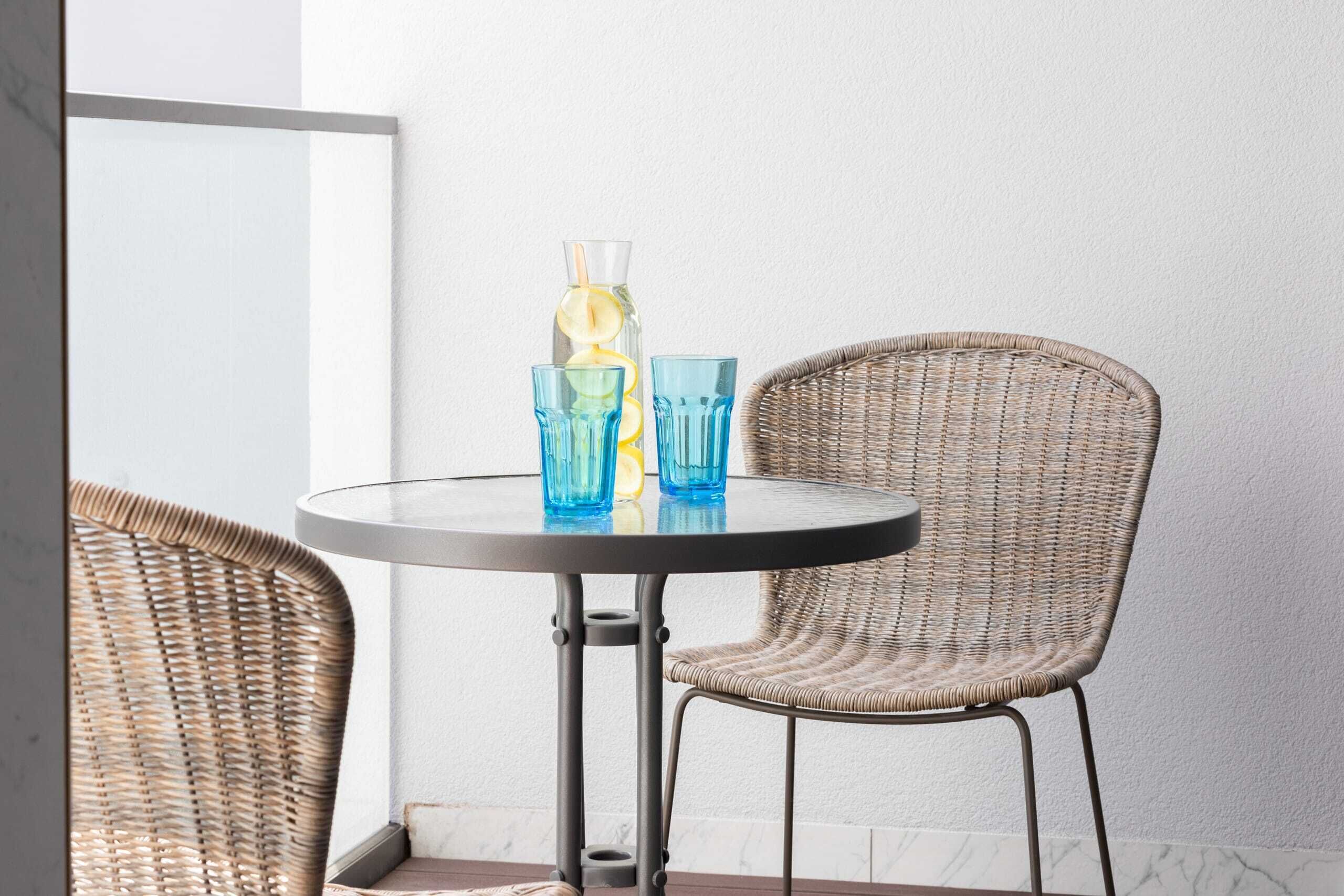 A round table with two blue glasses and a glass bottle with a straw, accompanied by wicker chairs.