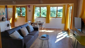 A cozy living room with a sofa, a coffee table, and a TV. There is a dining area with a table and chairs, and a kitchen area with a washing machine.
