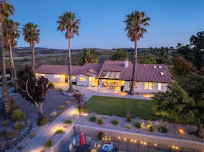 A two-story house with a patio, surrounded by palm trees and a landscaped garden.