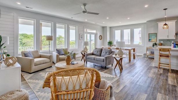 A spacious living room with large windows, a ceiling fan, and a kitchen area with white cabinets and a hanging light fixture.