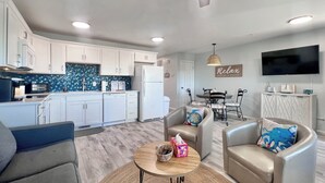 A modern living room with a sofa, armchairs, and a coffee table. There is a kitchen area with white cabinets and a tiled backsplash. A dining table with chairs is visible in the background.