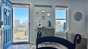 A room with a blue door, a wooden floor, a desk, a chair, a small cabinet, and two windows with a view of the outside.