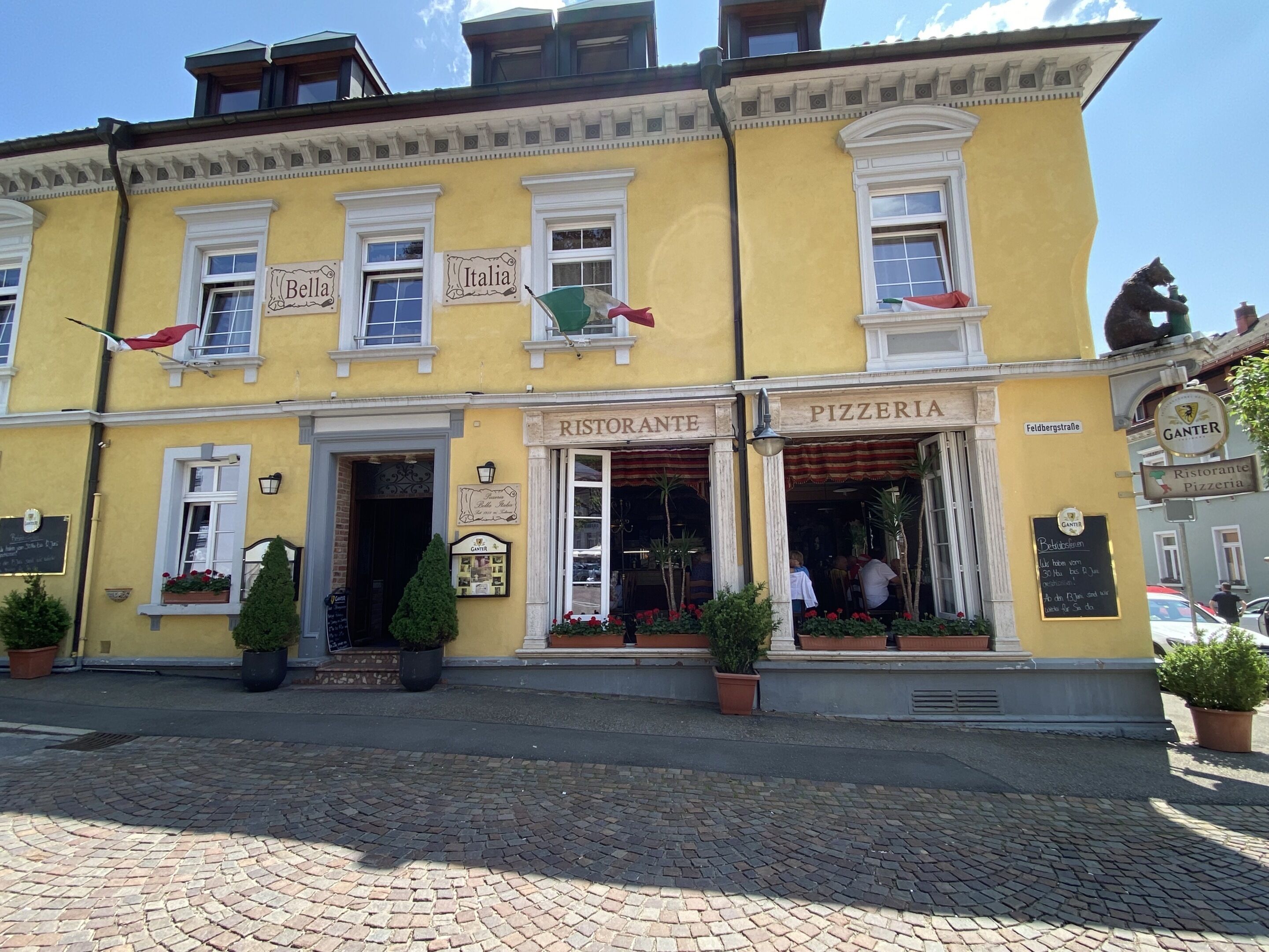Un bâtiment jaune dont le rez-de-chaussée abrite un restaurant dont l’enseigne indique « Ristorante » et « Pizzeria ». Des plantes en pot sont disposées devant l’entrée.