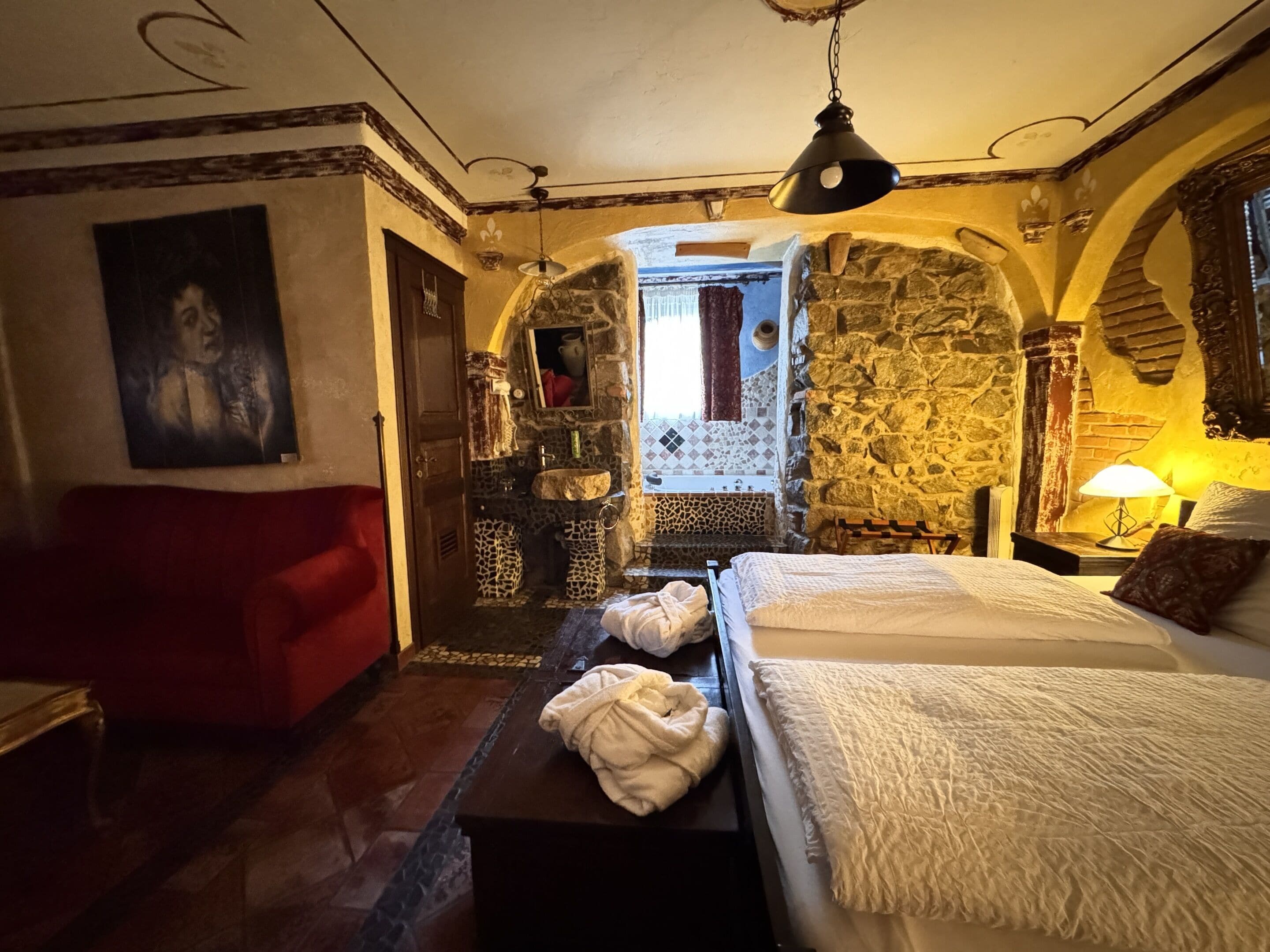 A cozy room with a bed, a red sofa, a stone wall, and a bathroom with a stone sink and mirror.