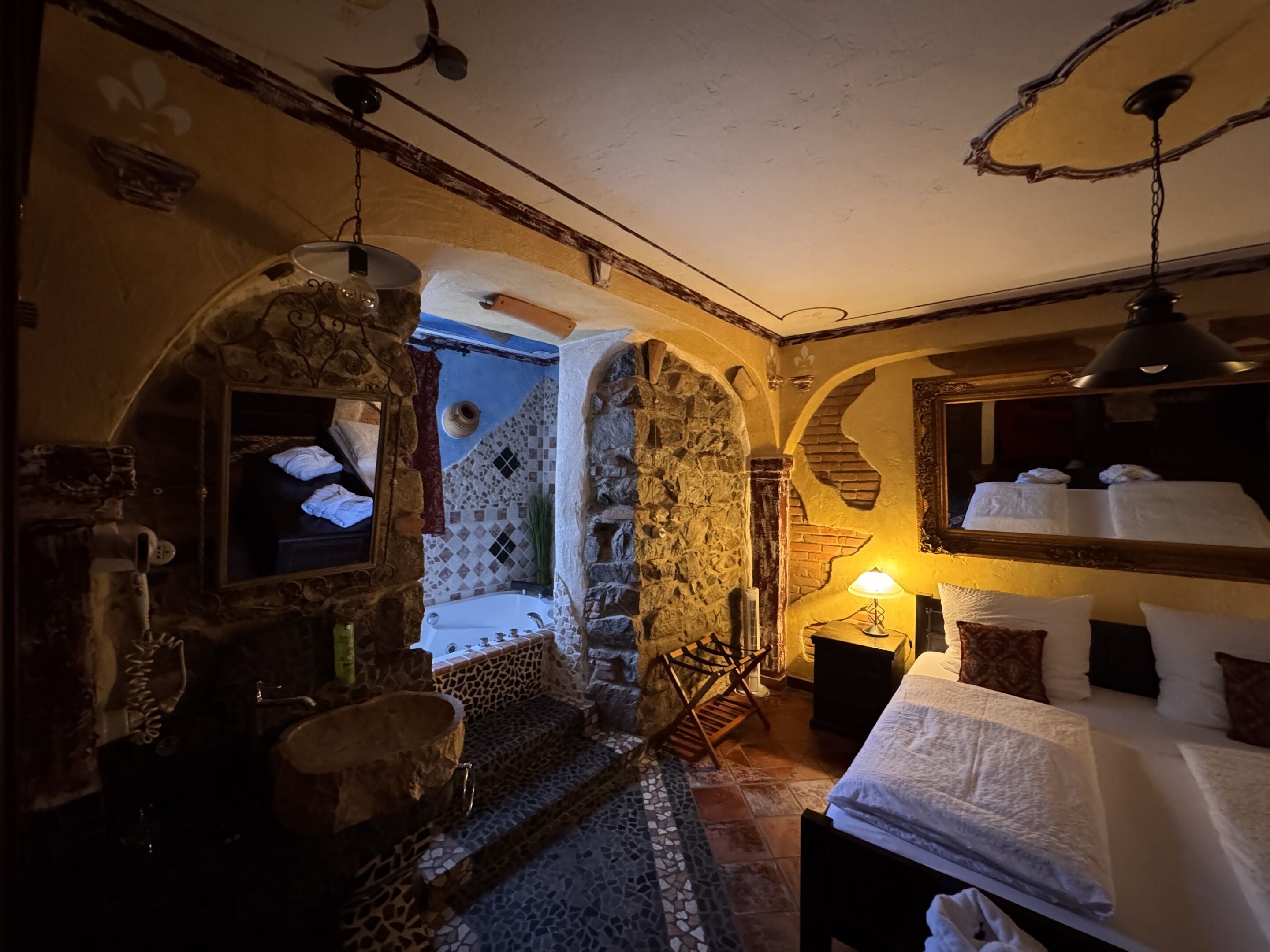 A rustic room with a stone wall, a wooden bench, a bed with white linens, and a hanging lamp.