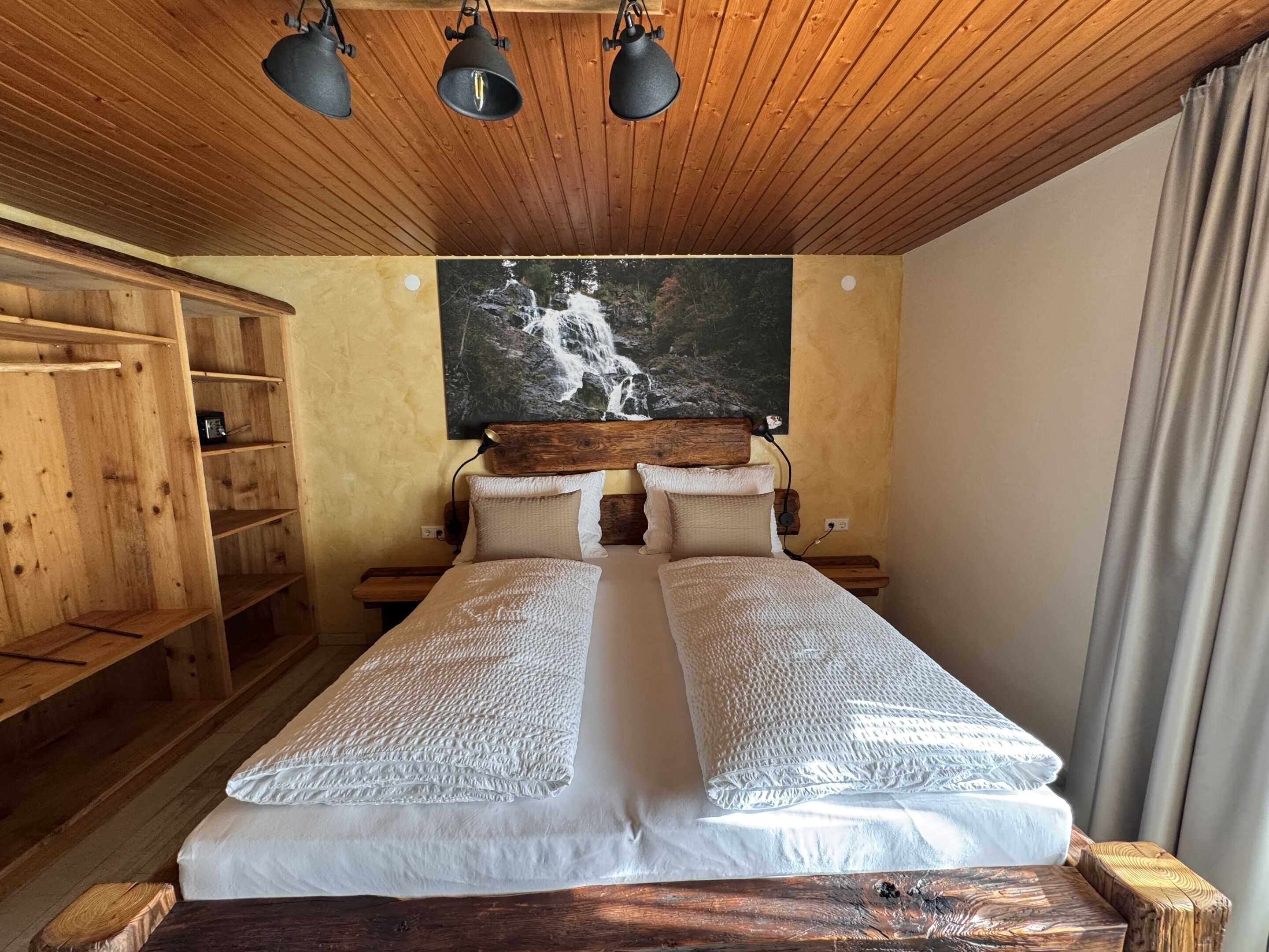 Una cama doble con cabecera de madera, una mesita de noche, una estantería y un cuadro de una cascada en la pared.