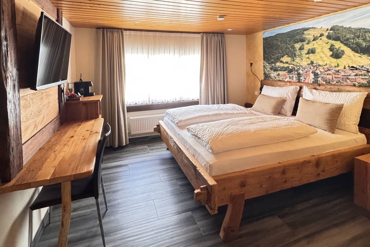 Un dormitorio con techo de madera, una cama con ropa blanca, un escritorio con silla, un televisor y una ventana con vista a un paisaje.