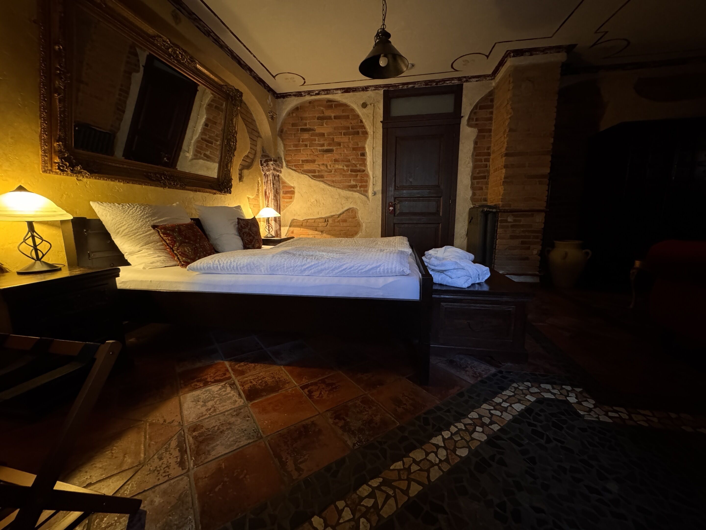Una habitación con poca luz, una cama, una mesita de noche, una lámpara y un espejo grande.