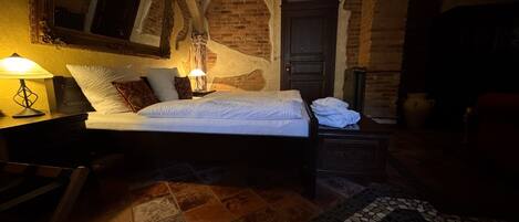 Una habitación con poca luz, una cama, una mesita de noche, una lámpara y un espejo grande.