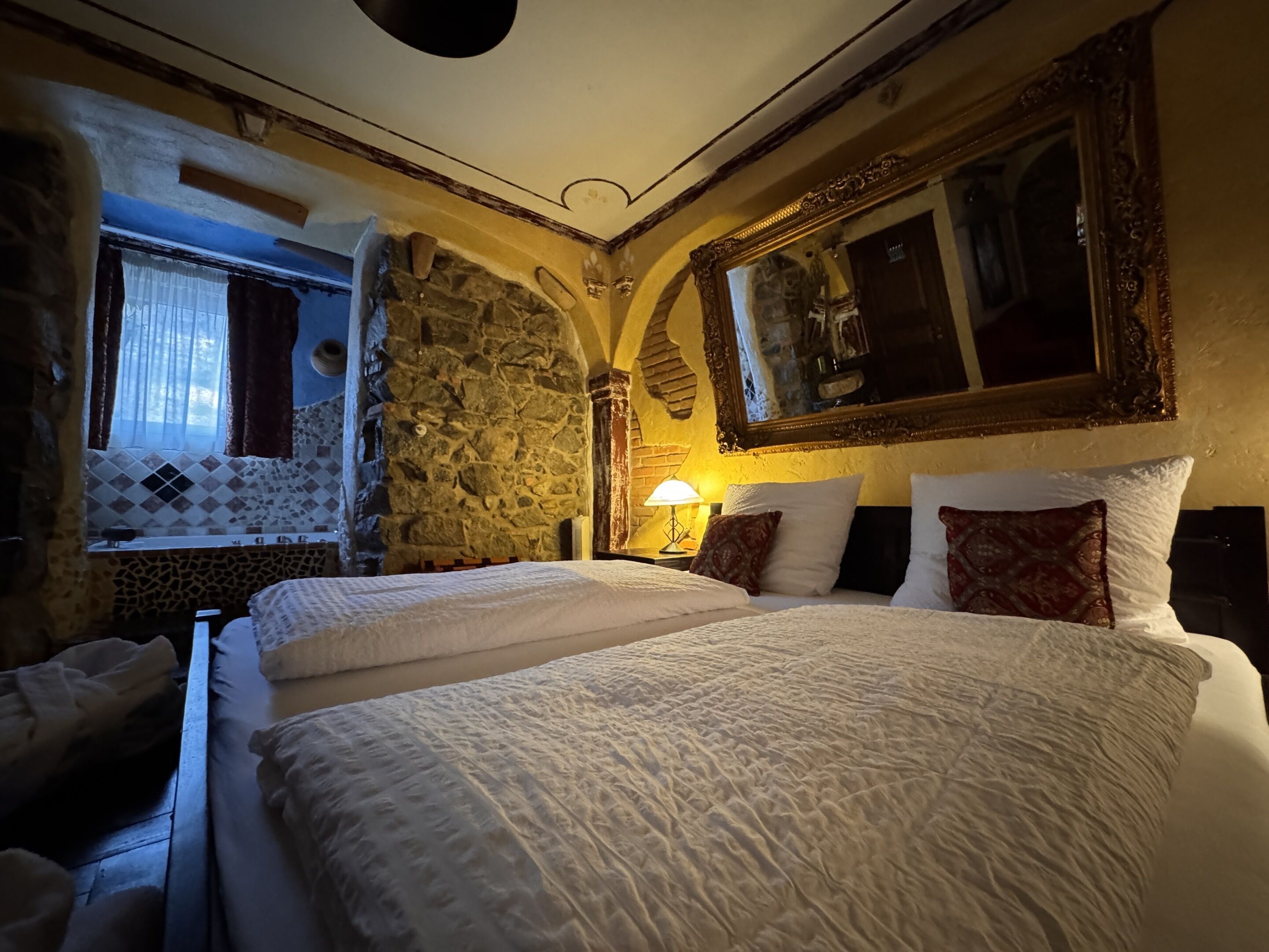 A hotel room with a stone wall, a large bed with white bedding, a mirror with an ornate frame, and a window with curtains.