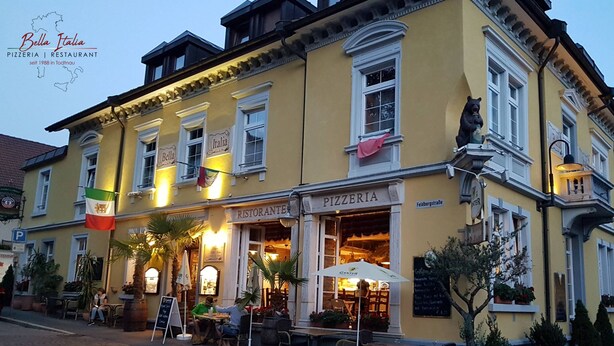 Een geel gebouw met een bord waarop 'Pizzeria' staat, zitplaatsen buiten en een vlag.