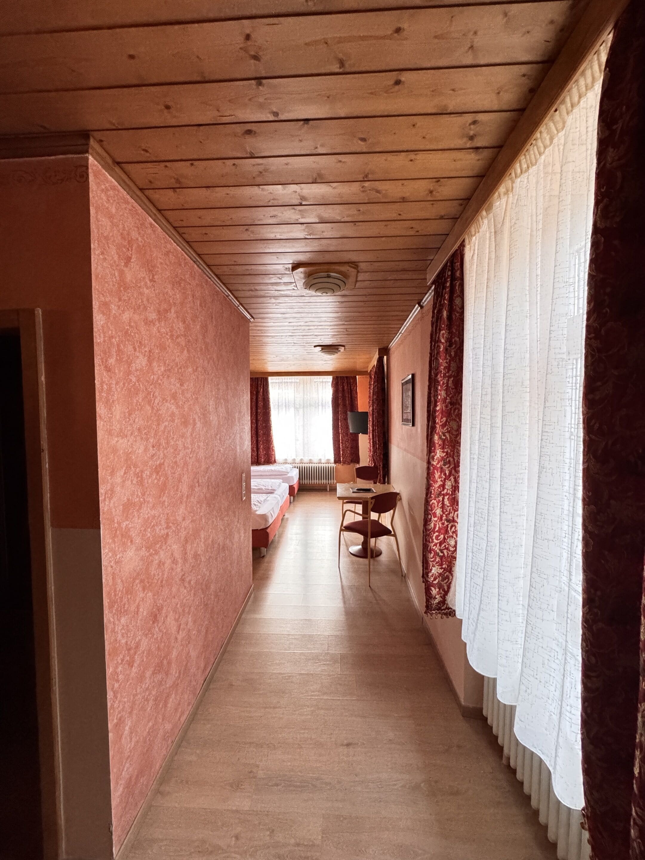 A narrow hallway with wooden ceilings and walls, leading to a room with a bed and a table.
