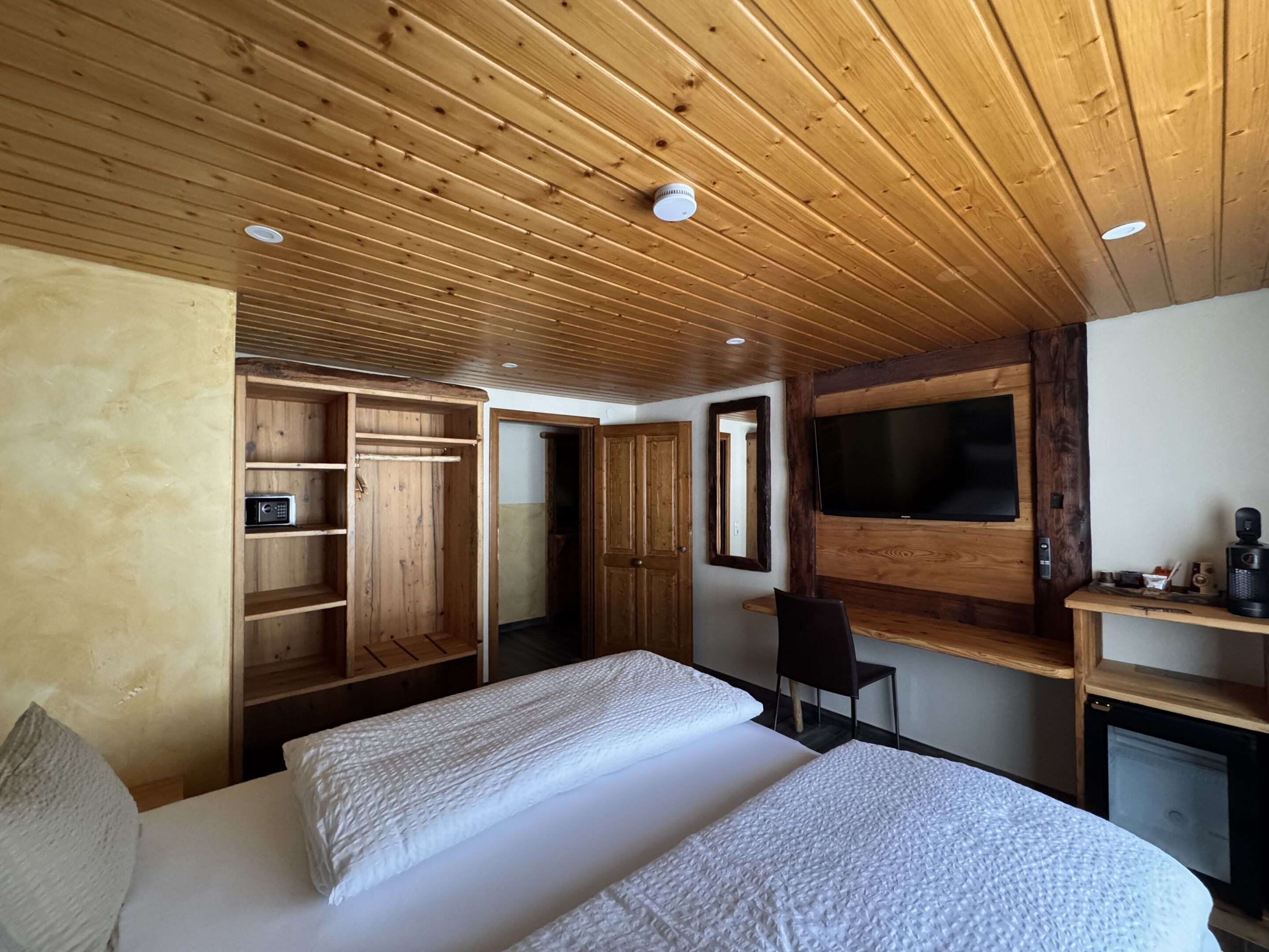 Un dormitorio con techo de madera, una cama con ropa blanca, un televisor montado en la pared y un escritorio de madera con una silla.