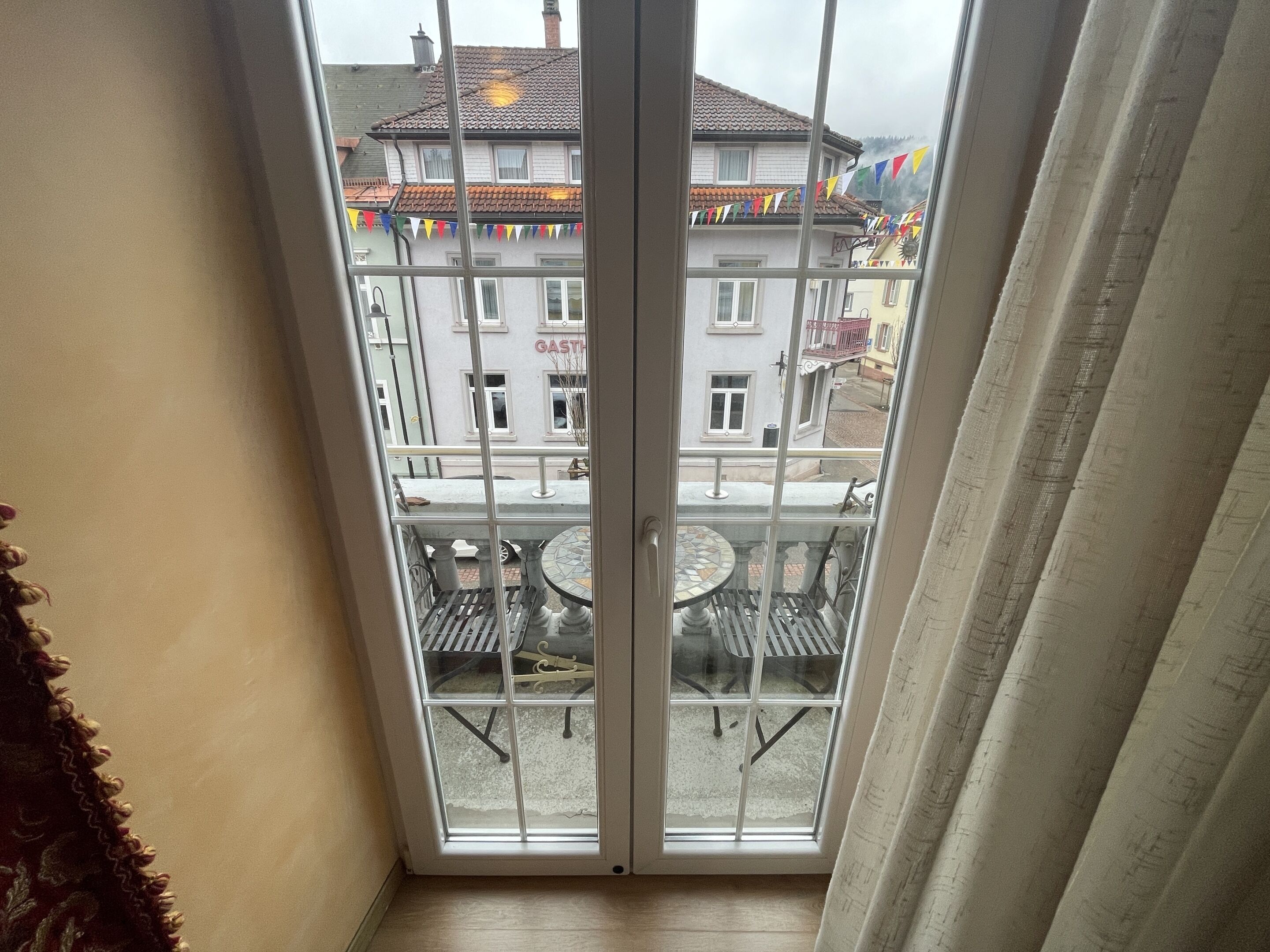 Ein Balkon mit Tisch und Stühlen, Blick auf ein Gebäude mit der Aufschrift 'GASTHAUS' und ein Fenster mit halb heruntergezogenem Vorhang.