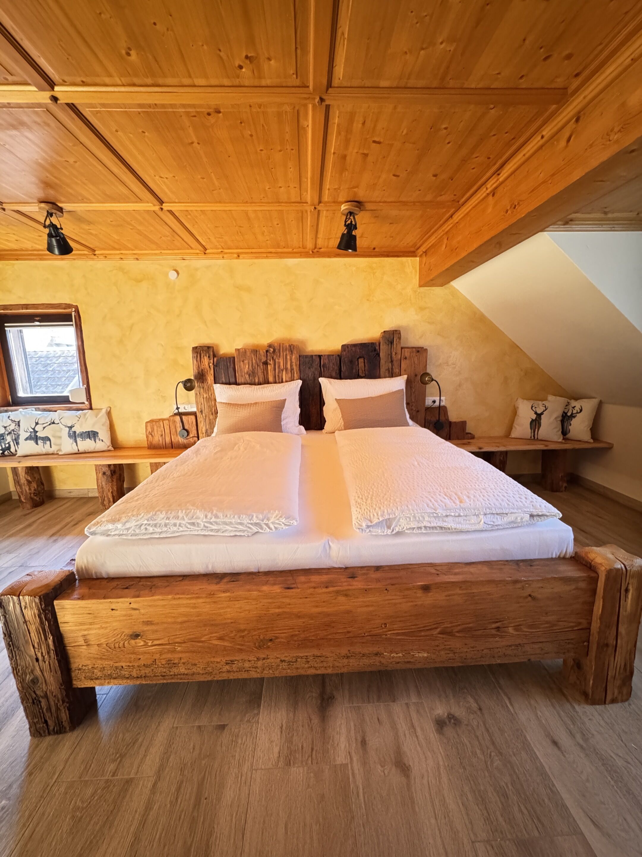 Ein hölzernes Bett mit einem Blockbohlen-Kopfstück, eine Holzbank mit Kissen und eine geneigte Decke.