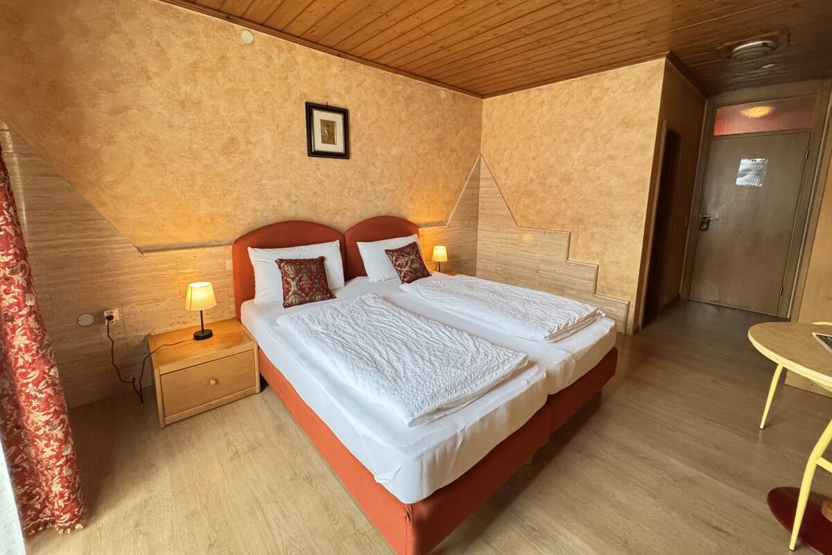Una cama doble con ropa de cama blanca, dos lámparas de noche, un cabecero de madera, un cuadro enmarcado en la pared y suelo de madera.