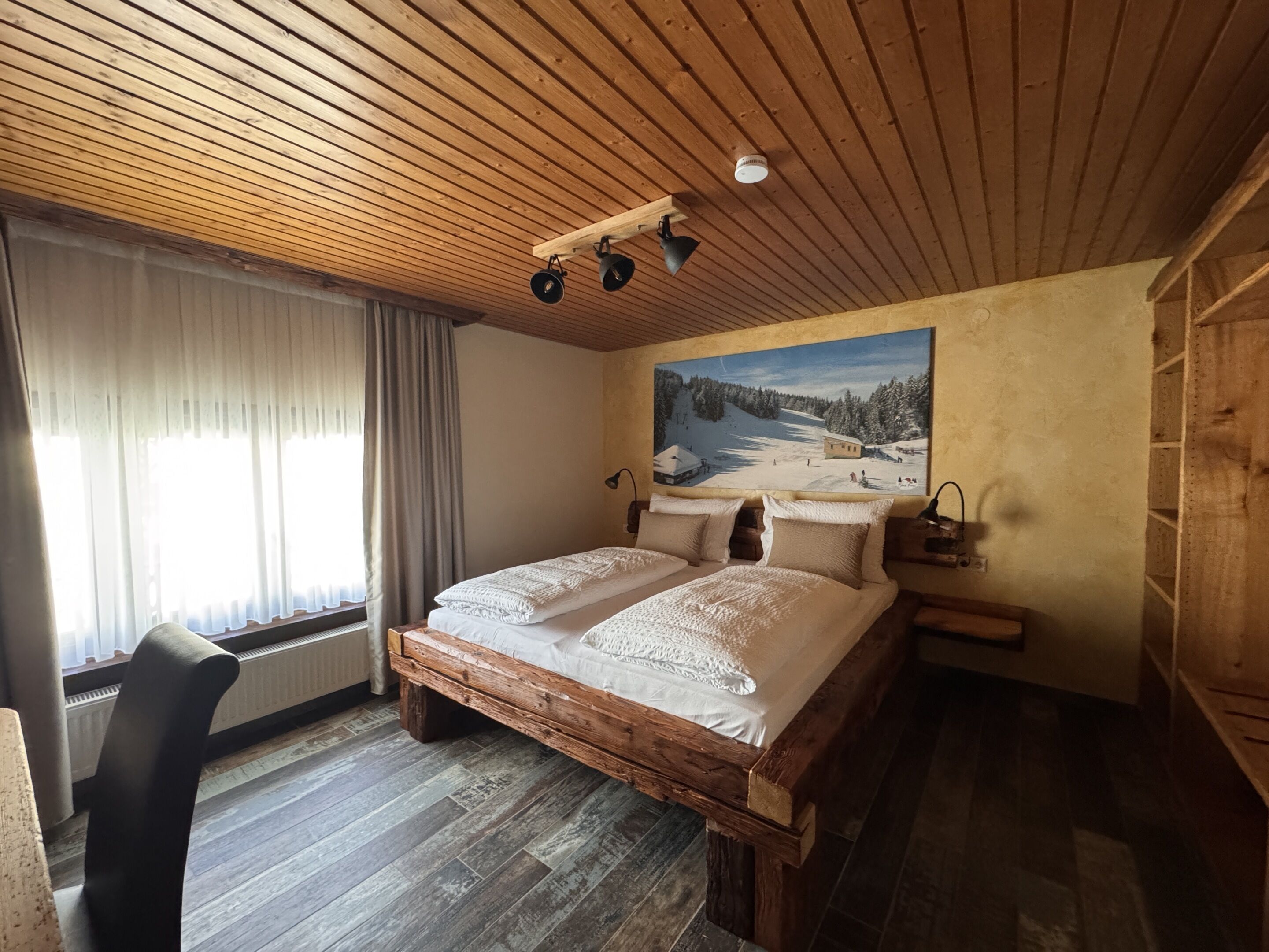 Techo de madera, una cama con sábanas blancas, un cuadro de un paisaje nevado y una silla.