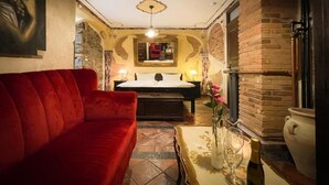 Een knus ingerichte kamer met een rode bank, een bed met nachtkastje, een vaas met bloemen en een bakstenen muur.