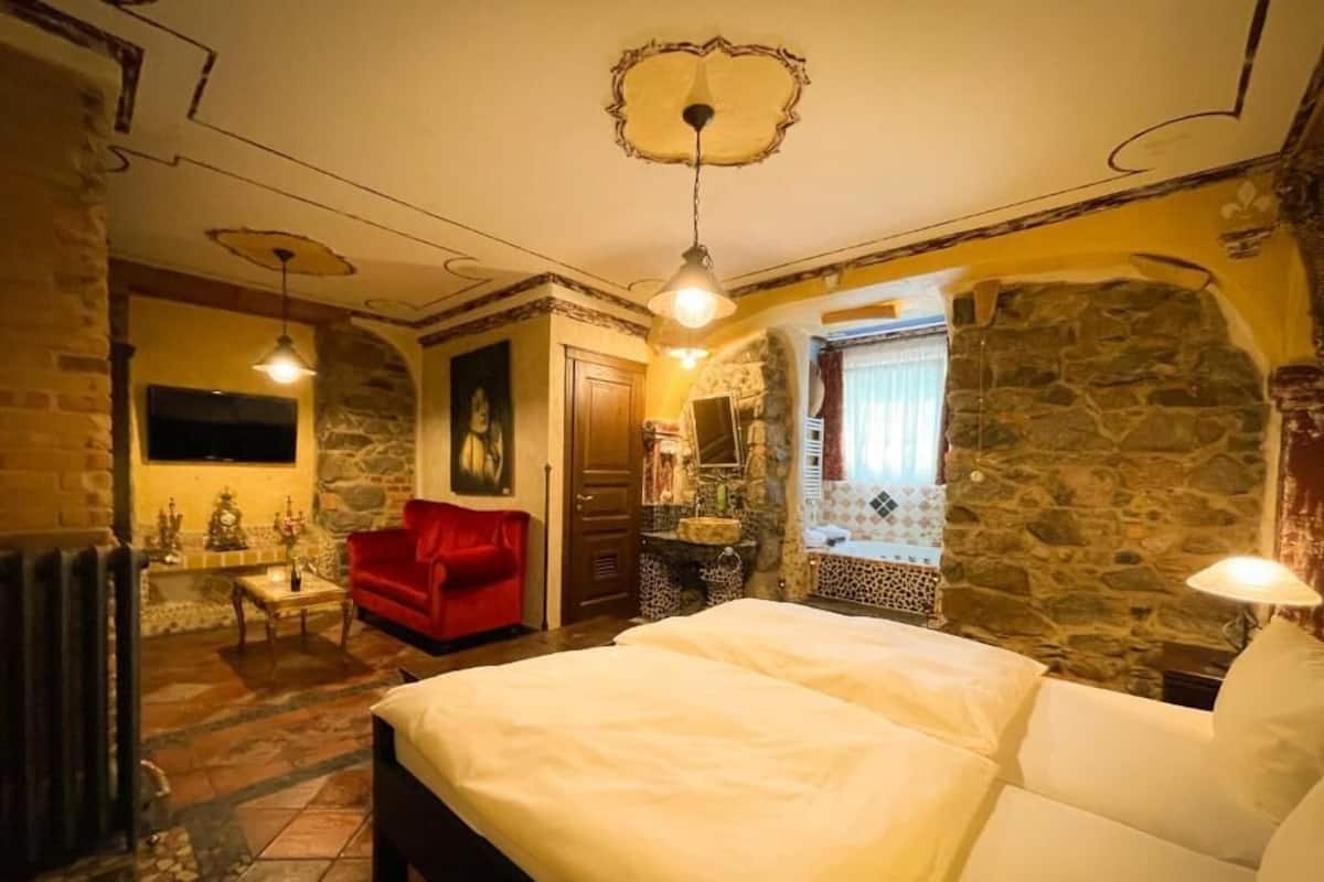 Un dormitorio con una cama grande, un sillón rojo, una pared de piedra y una ventana con cortinas.