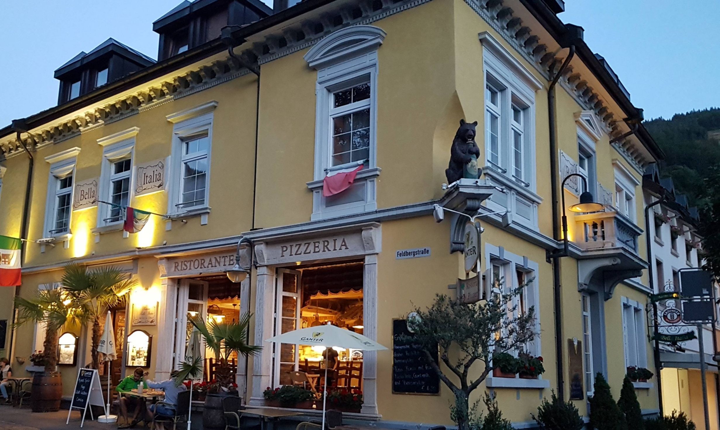 A yellow building with a 'Pizzeria' sign, outdoor seating, and a flag.