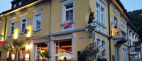 A yellow building with a 'Pizzeria' sign, outdoor seating, and a flag.