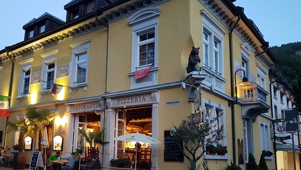 A yellow building with a 'Pizzeria' sign, outdoor seating, and a flag. - Black Forest Hotel (Todtnau)