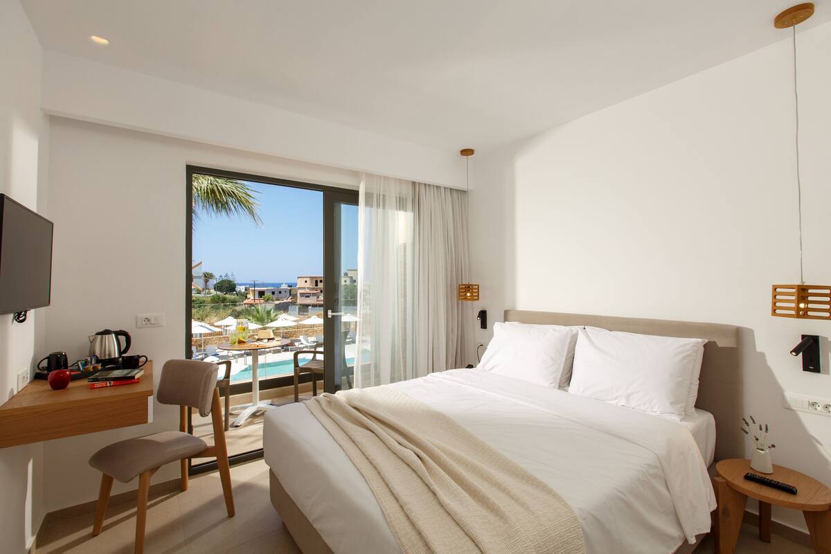 Habitación de hotel con cama, escritorio y silla, televisor y vistas a una piscina y edificios.