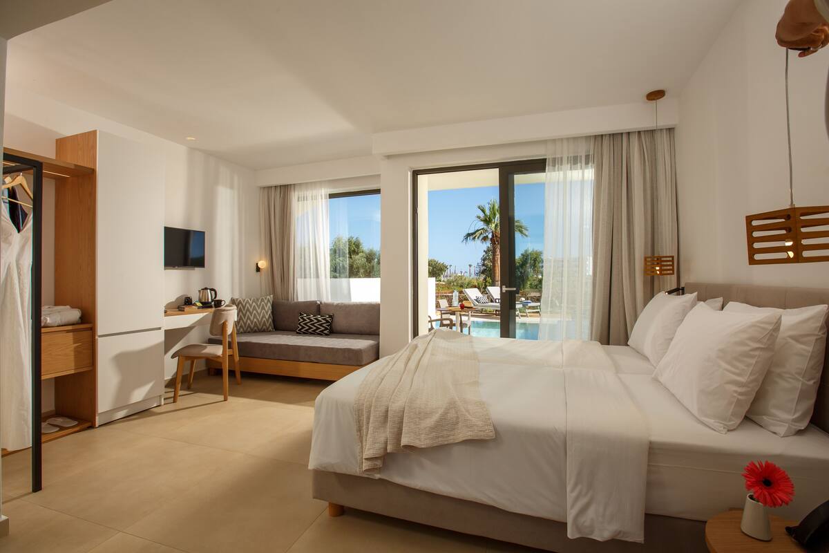 Habitación de hotel moderna con una cama grande, un escritorio, un sofá y vistas a una piscina y palmeras.