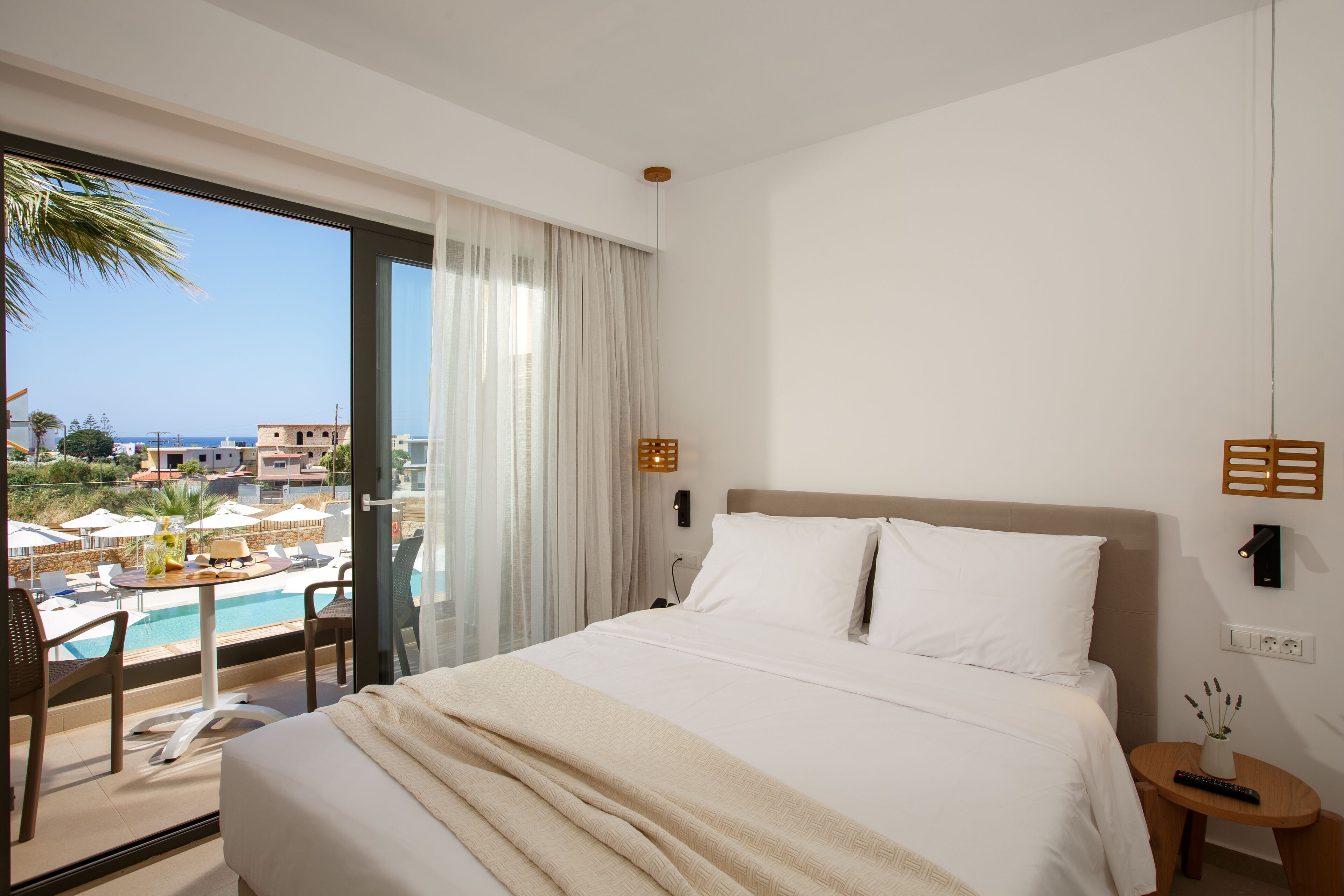 A hotel room with a large bed, a view of a pool and buildings, and a balcony with chairs.