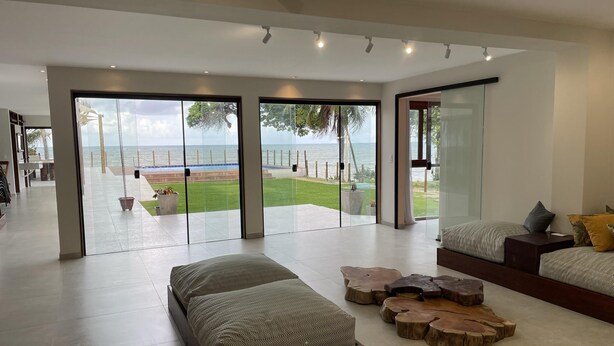 A spacious living area with a large sliding glass door leading to a balcony with a view of the ocean. There are two seating areas with cushions and a coffee table. - Front sea Mansion With Pool Maceio Paripueira (Paripueira)