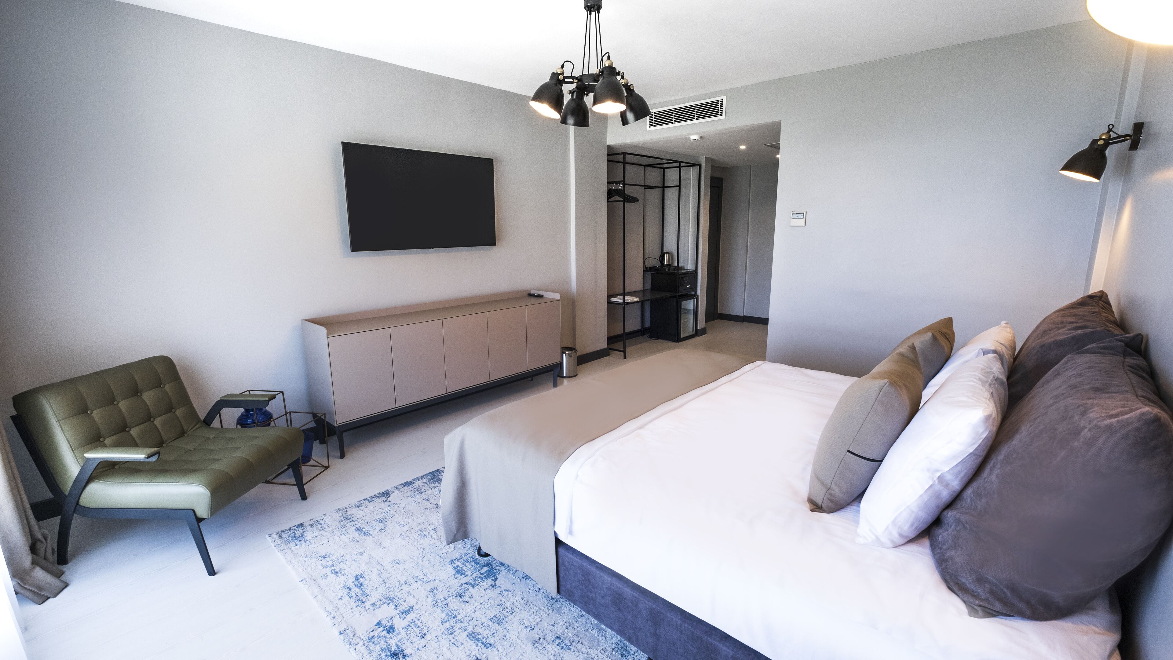 Un dormitorio moderno con una cama grande, una zona de estar con una silla, un televisor montado en la pared y un espejo que refleja la habitación.