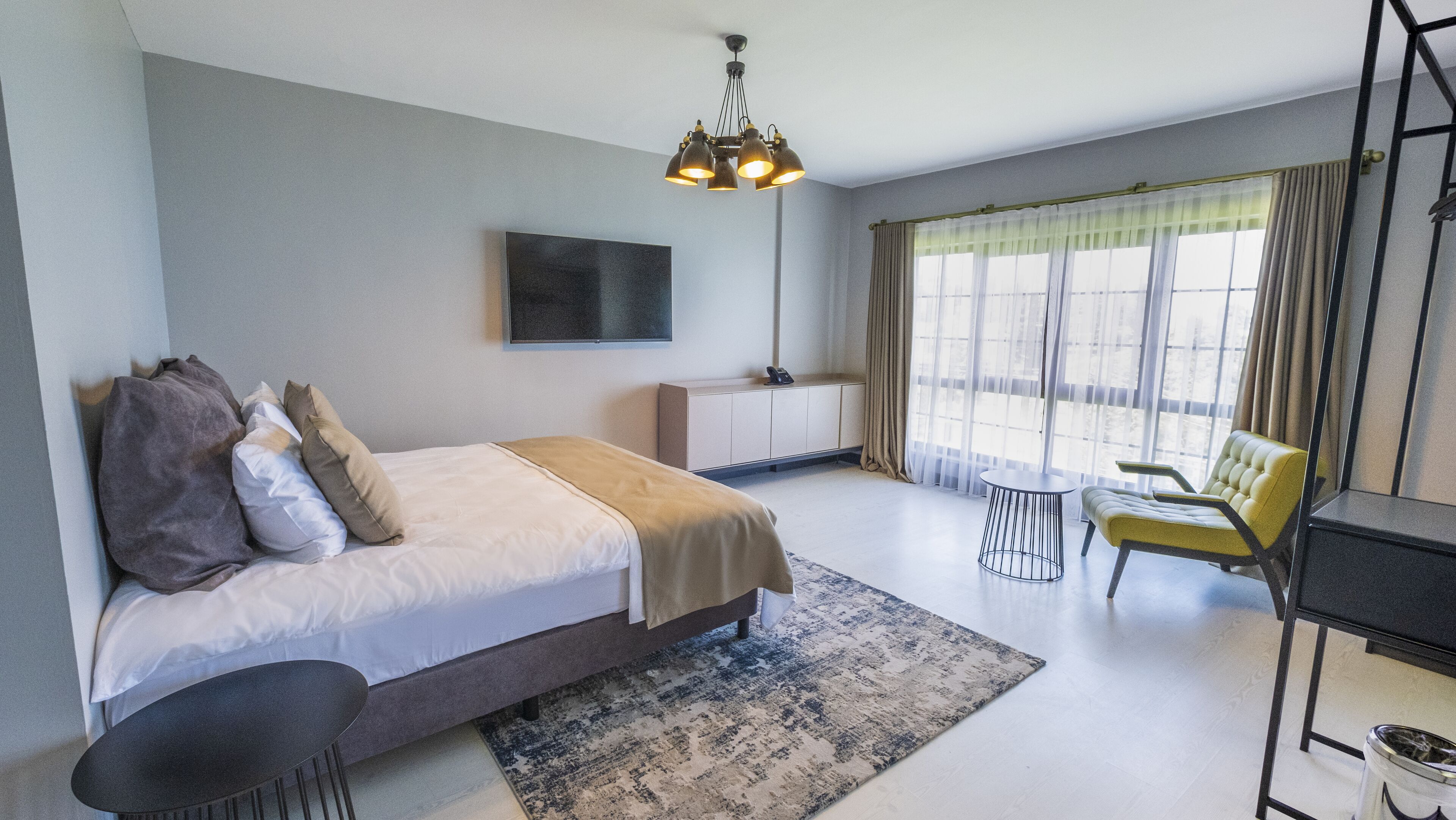 A modern hotel room with a large bed, a desk, a chair, a TV, and a large window with curtains.