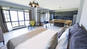 A modern hotel room with a large bed, a seating area with a chair, a small table, and a kitchenette in the background.