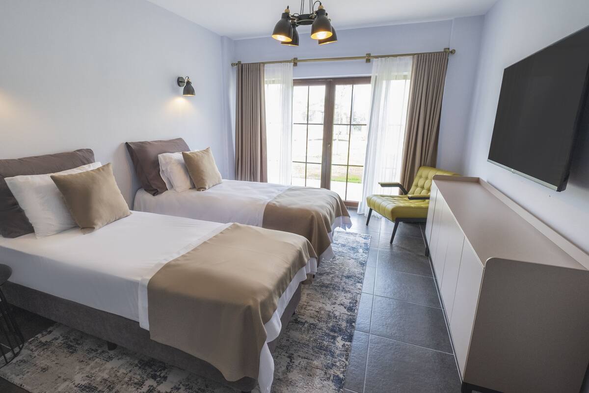 Ein Hotelzimmer mit zwei Betten, einem Fernseher, einem Sessel und einem Fenster mit Vorhängen.