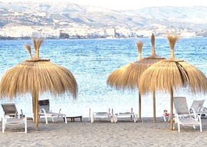 A beach with thatched umbrellas and lounge chairs facing a body of water with a cityscape in the background. - Fabulous Apartment close to La Duquesa Marina, beaches, shops, and restaurants (Manilva,)