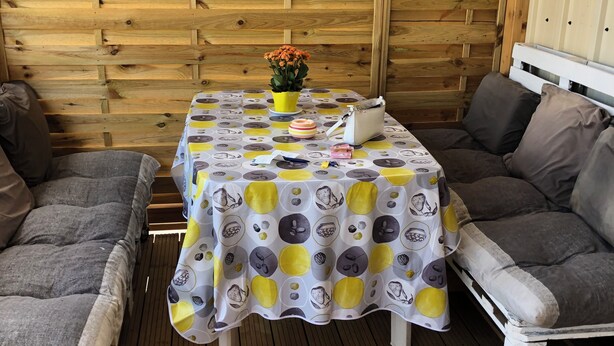 A wooden deck with a table covered by a patterned tablecloth, two cushioned chairs, a potted plant, and a handbag on the table.