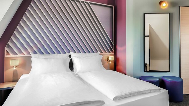 A modern hotel room with a bed, bedside lamps, a mirror, and a unique slatted headboard design. - B&B HOTEL Gießen-City (Giessen)
