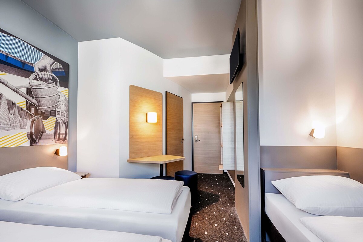 Habitación de hotel con dos camas, un mural en la pared y un pasillo que lleva a una puerta.
