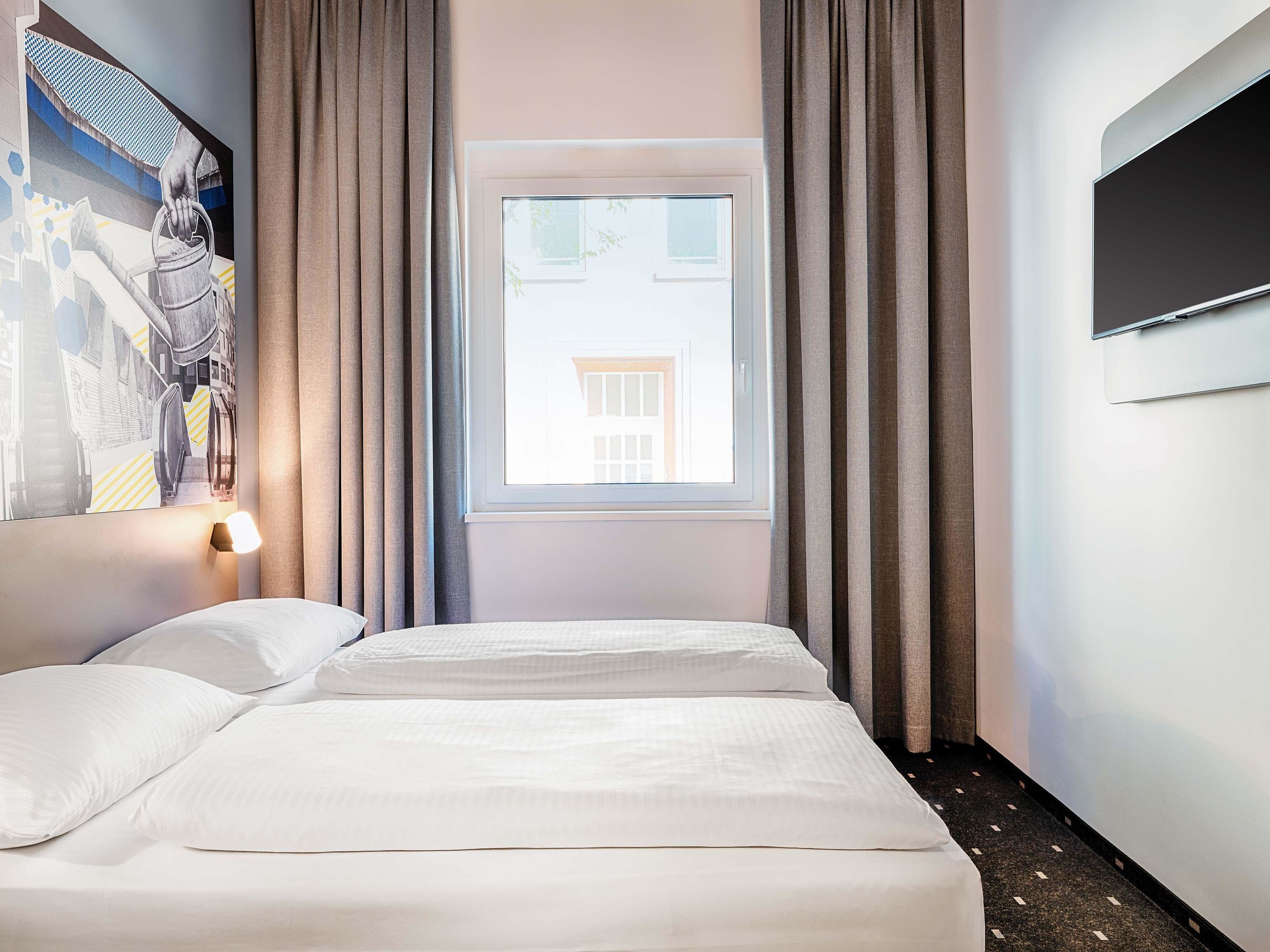 A hotel room with a bed, a window with curtains, a wall-mounted TV, and a painting on the wall.
