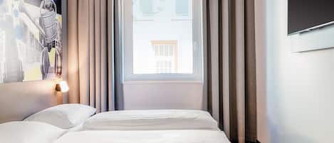 A hotel room with a bed, a window with curtains, a wall-mounted TV, and a painting on the wall.