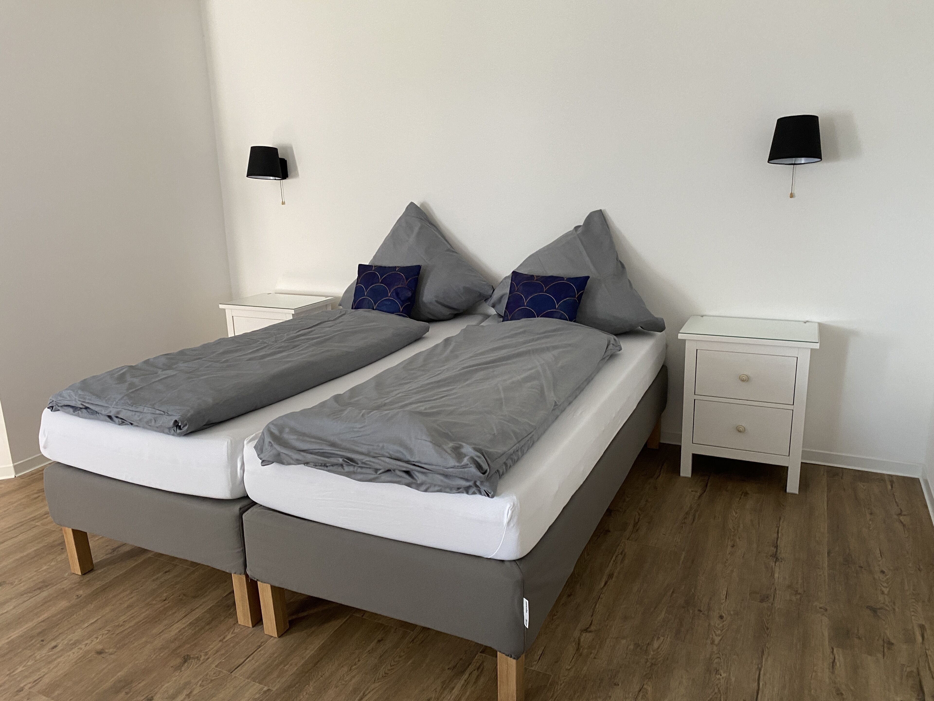 Una cama doble con ropa de cama gris, dos mesitas de noche y dos lámparas de pared.
