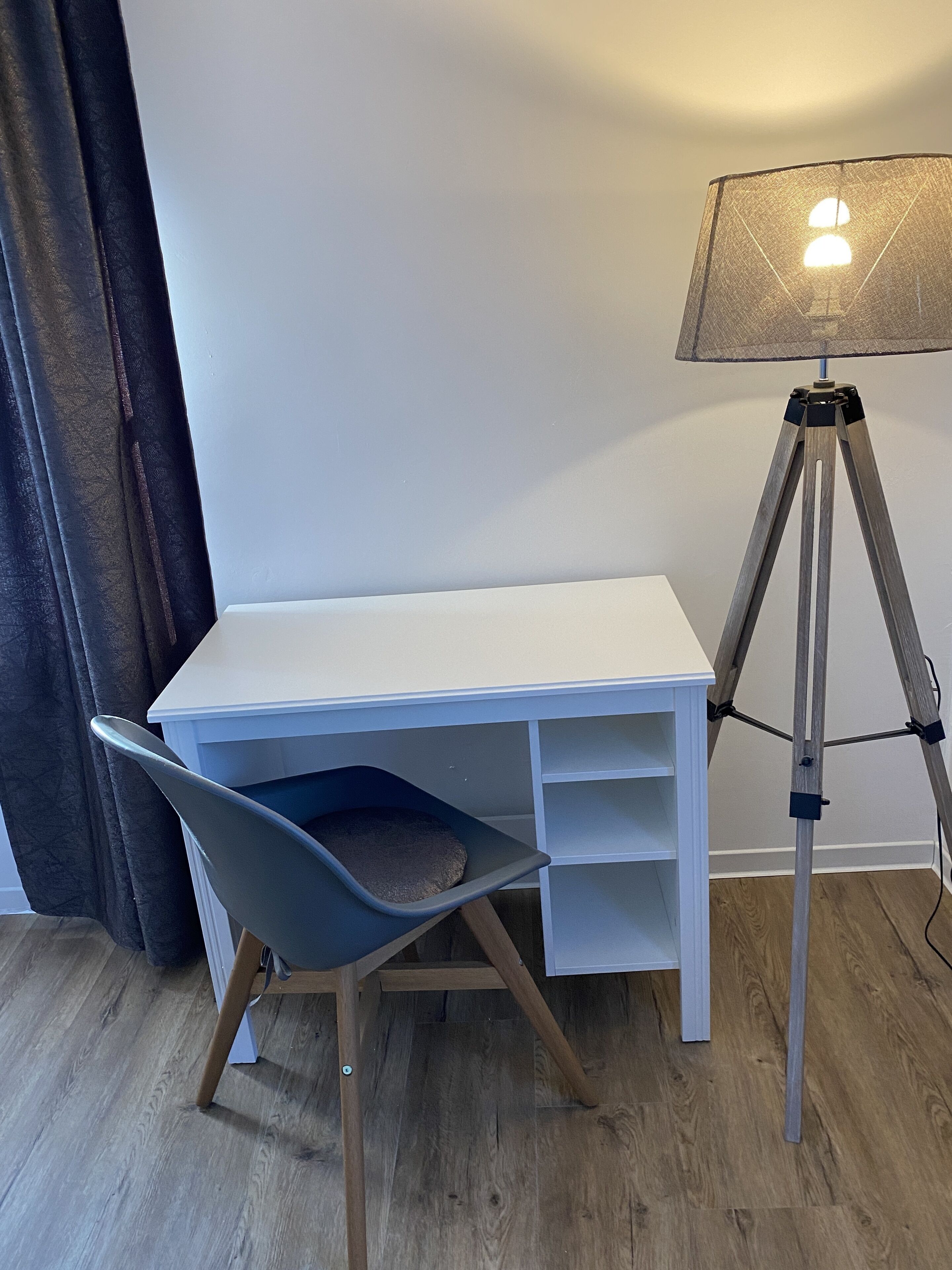 Un bureau blanc avec une chaise et une lampe sur pied, dans une pièce dont le sol est recouvert de parquet et dont un des murs est uni.