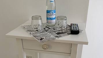 Ein kleiner weißer Tisch mit einer IsarPerle-Flasche, zwei Gläsern, einer Fernbedienung und einem Buch auf einer Unterlage.