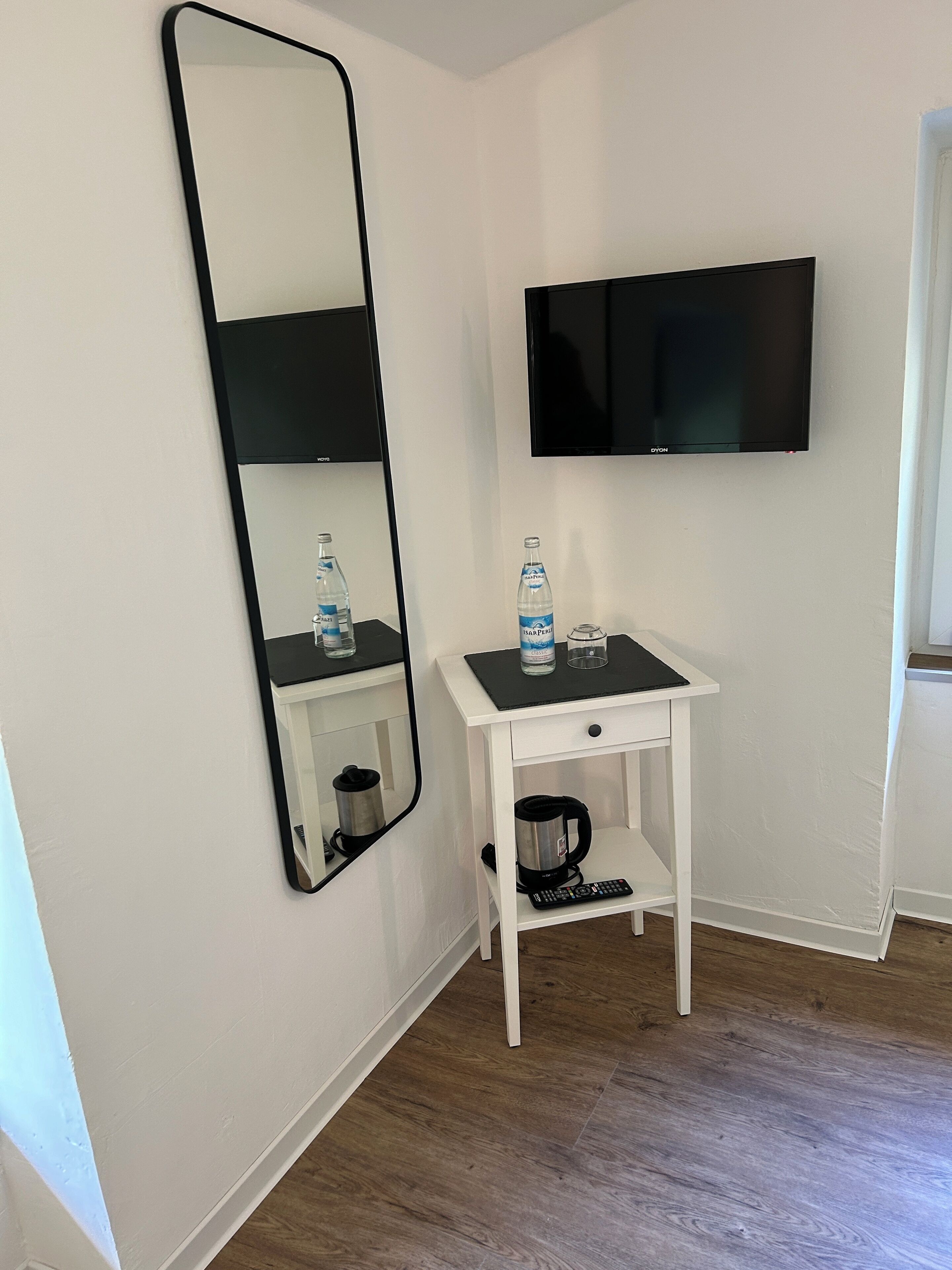 Una habitación pequeña y blanca con piso de madera, un escritorio de madera con cubierta negra, un espejo con marco negro, un televisor de pantalla plana y una cafetera.