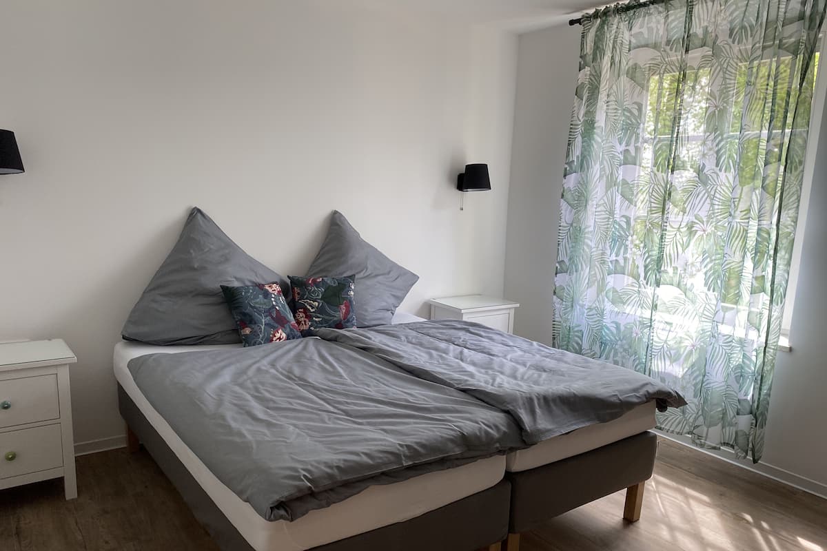 Cama bem arrumada com roupa de cama cinza e almofadas decorativas, mesa de cabeceira com abajur e janela com cortina estampada com folhas.