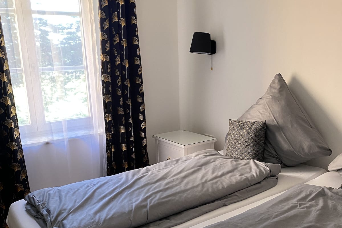 Quarto com duas camas, mesa de cabeceira, abajur e janela com cortinas.